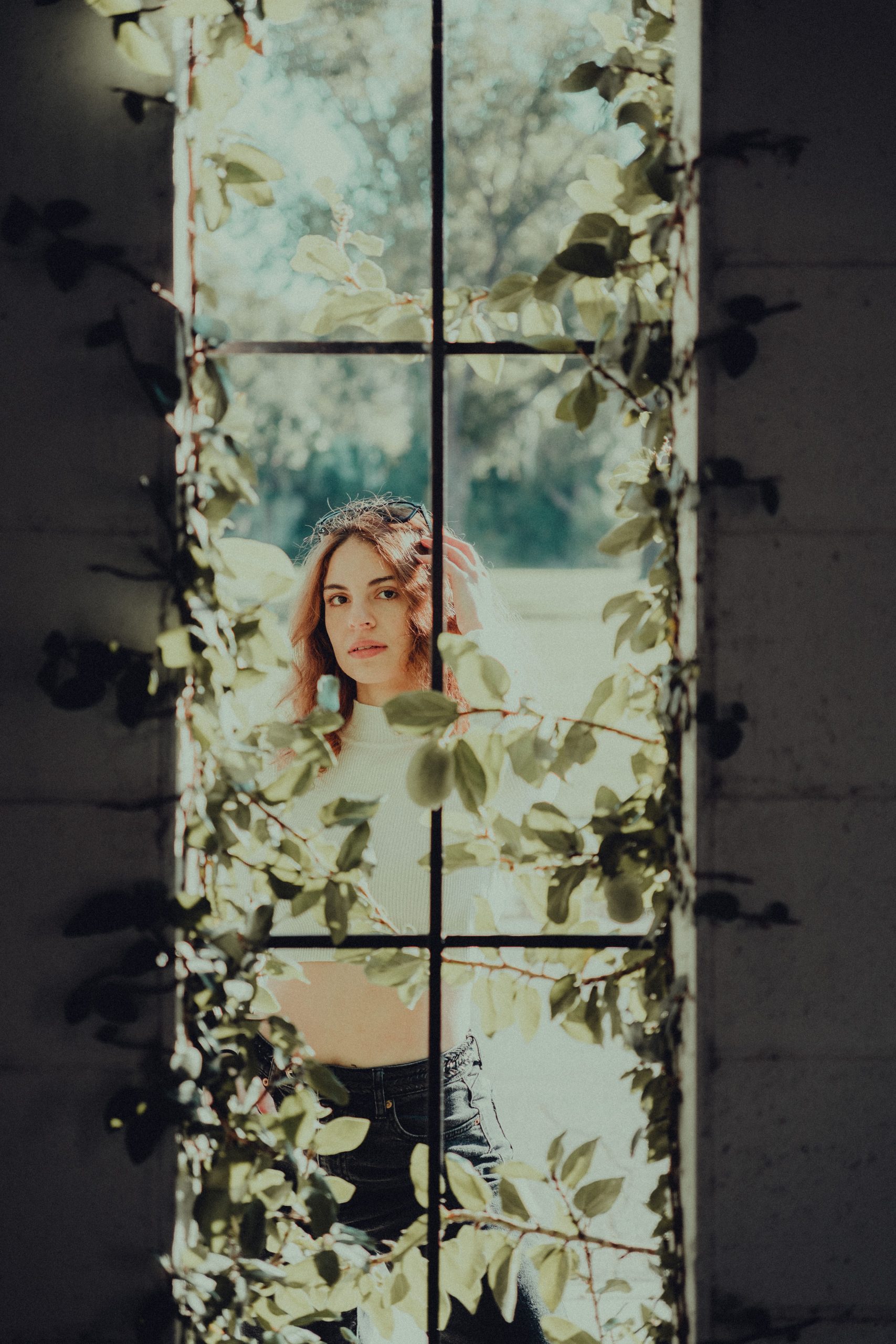 A woman stands behind a window adorned with sunlit vines, creating a serene and natural frame.