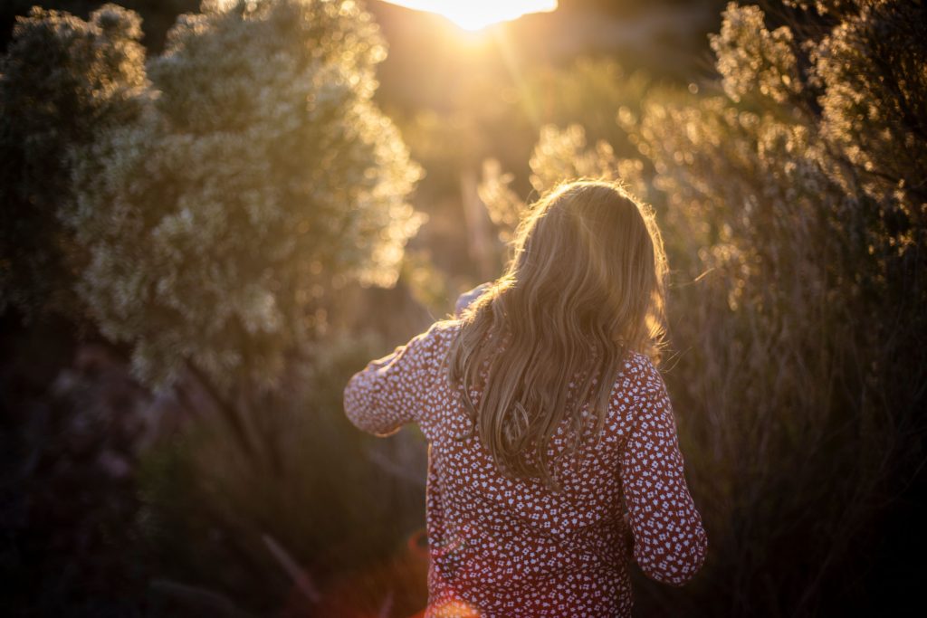 A woman with blonde hair walks through a sunlit field at sunset, capturing tranquility.