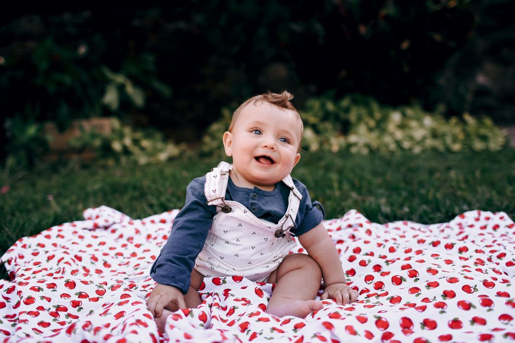 A happy baby sitting on a cherry-printed blanket outdoors, enjoying a sunny day.